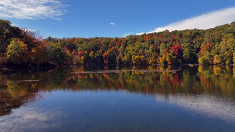 An-aerial-view-over-a-reflective-lake,-surrounded-by-colorful-trees-during-the-fall-foliage-in-upstate-NY