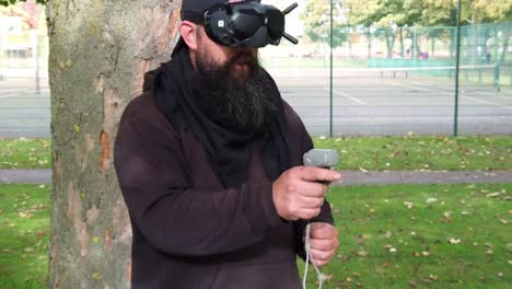 Bärtiger-Mann-Mit-Augmented-Reality-FPV-Brille,-Der-Drohne-Im-Park-Mit-Bewegungs-Joystick-Steuert