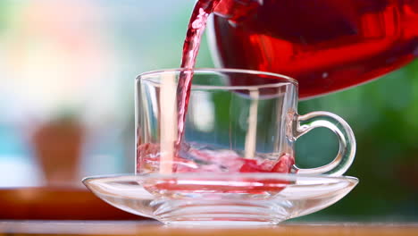 Pouring-red-herbal-tea-out-of-transparent-tea-pot-into-glass-with-herbal-leaves,-lemon-and-ice-cubes-with-green-nature-background