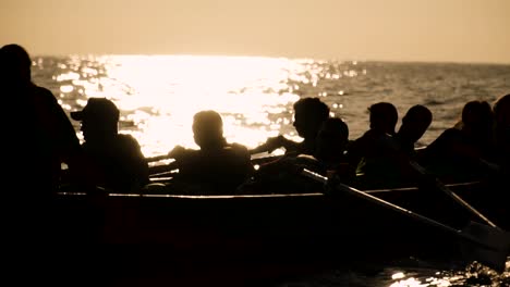 Silhouette-of-team-of-people-rowing-at-sea-in-front-of-bright-sunset-or-sunrise