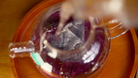 Top-view-of-pouring-hot-water-into-transparent-glass-tea-pot-with-tea-bag-and-herbal-leaves