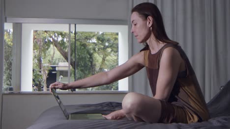 a-female-model-sits-on-bed-and-opens-her-laptop-computer-to-work,-slow-motion-close-up