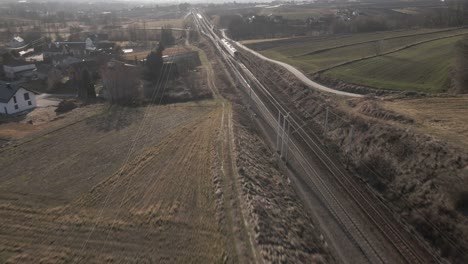 Aerial-view-of-passanger-train-on-a-sunny-day-during-the-golden-hour,-next-to-agricultural-fields-and-villages---concept-of-comfortable-passanger-transportation