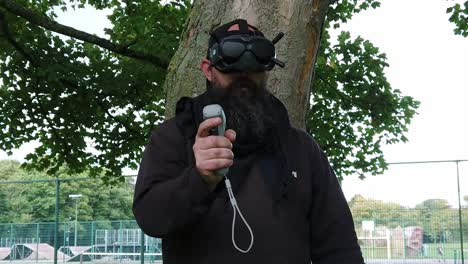 Bearded-male-wearing-digital-FPV-goggles-controlling-drone-in-park-with-motion-joystick