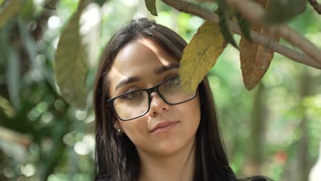 Beautiful-Green-Eyed-Latina-Woman-with-Glasses-Looking-into-Camera-from-Behind-Branch-in-Park