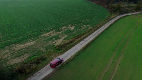 Aerial-drone-forward-moving-shot-over-red-car-driving-through-road-among-agriculture-fields-along-green-countryside-during-daytime