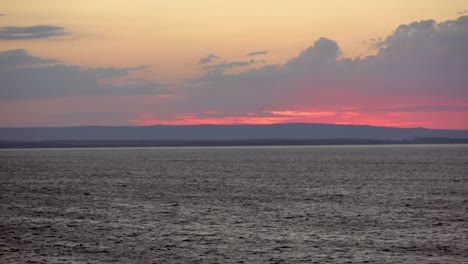 View-from-sea-of-sun-setting-behind-clouds-and-hills-near-Sydney,-Nova-Scotia-with-beacon-flashing-on-shore