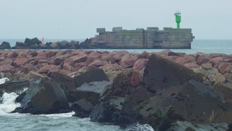 Rolling-waves-crashing-along-pier-wall-and-stones,-overcast-autumn-day,-distant-green-port-navigation-light-in-background,-climate-changes,-medium-shot