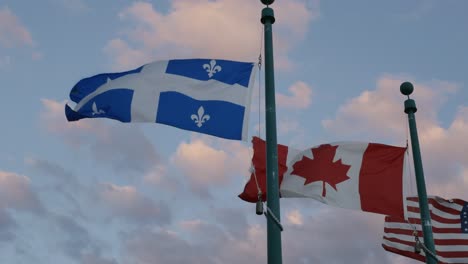 Flags-Of-Quebec,-Canada,-And-America-Waving-Against-Cloudy-Sunset-Sky-In-Magog,-Canada
