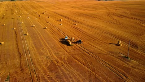 An-aerial-view-of-a-large-industrial-brown-field-with-a-tractor-collecting-hay-bales,-zooming-in