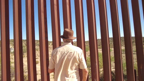 Focus-on-man-with-hat-looking-through-US-Mexico-border-fence,-then-reversing-and-showing-barbed-wire-on-top