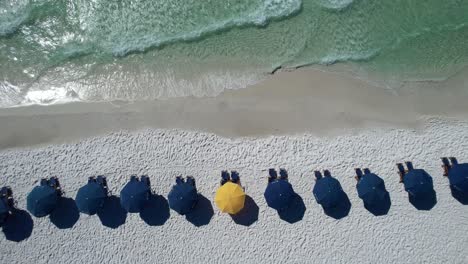 Blue-umbrellas-on-Destin-Beach-gulf-coast-panhandle-Florida-white-sandy-beach-clear-emerald-waters-fly-over-with-drone-aerial-view-bright-sunny-day