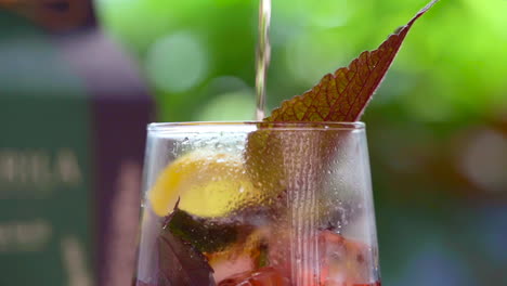 Pouring-red-herbal-tea-out-of-transparent-tea-pot-into-glass-with-herbal-leaves,-lemon-and-ice-cubes-with-green-nature-background