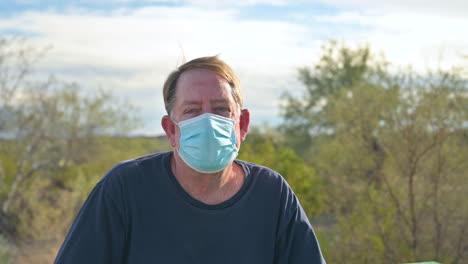 Senior-adult-man-outdoors-in-public-park-wearing-medical-face-mask,-looks-at-camera