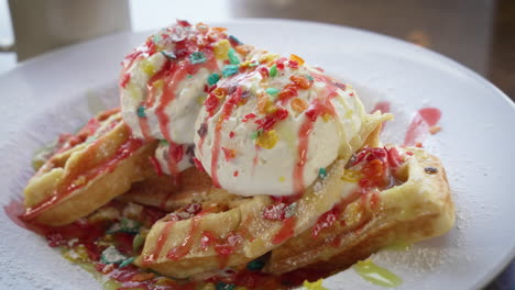 Confetti-Belgian-waffles-filled-and-topped-with-fruity-pebbles-cereal-and-ice-cream-drizzled-with-strawberry-and-maple-syrup,-close-up-slider-4K