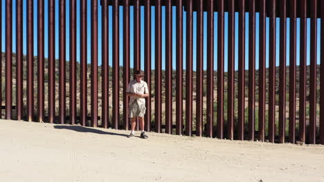 Drone-flies-towards-older-American-guy-in-hat-in-front-of-USA-Mexico-border-wall-in-California