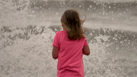 Wellen-Spritzen-Auf-Ein-Kleines-Mädchen-Hoch,-Das-Am-Strand-Steht-Und-Auf-Das-Meer-Blickt