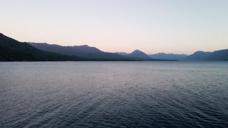 Dolly-out-of-a-lake-with-mountains-on-the-horizon-in-the-blue-hour-and-birds-flying-around---aerial