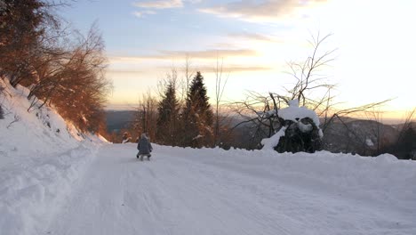 female-on-sled-carrying-a-baby-slowly-descends-down-snowy-road-into-golden-sunset