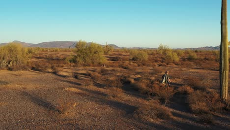 A-southwestern-desperado-man-wearing-a-poncho-walks-through-sagebrush-and-past-towering-Saguaro-Cacti-in-the-Sonoran-Desert-at-sunset