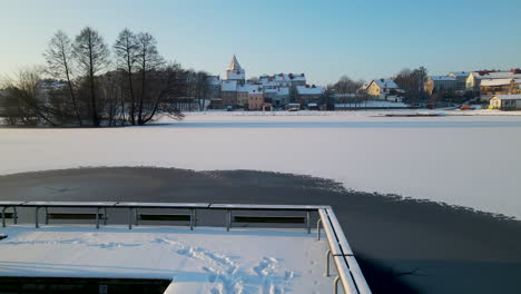 Schneebedeckte-Brücke-über-Einem-Zugefrorenen-See-Mit-Fußspuren-In-Einer-Kalten,-Verschneiten-Stadt
