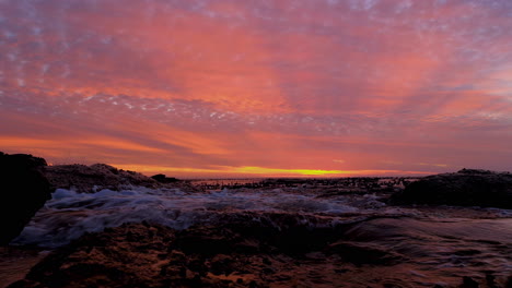 Gentle-waves-crash-onto-rocky-coastline,-vivid-orange-clouds-at-sunset