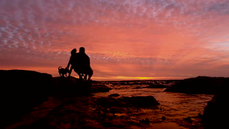 Father-shows-young-daughter-and-small-dog-vibrant-sunset-on-coastline,-silhouettes