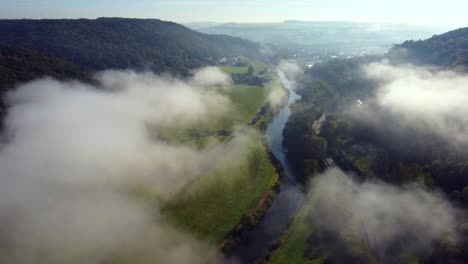 Morning-light-hits-the-valley-where-a-river-is-flowing-through,-while-clouds-are-moving-above-it-in-the-countryside-of-Luxembourg