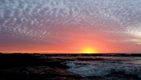Gentle-waves-crash-on-rocks,-clouds-glowing-orange-at-sunset-over-ocean