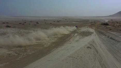 Aerial-View-Of-Dust-Trail-From-4x4-Driving-Across-Arid-Desert-Landscape-With-Mud-Volcano-In-Background