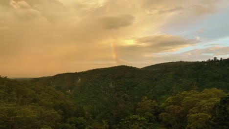 A-drone-flying-over-a-lush-forest-towards-rainbow-and-oncoming-rain-clouds
