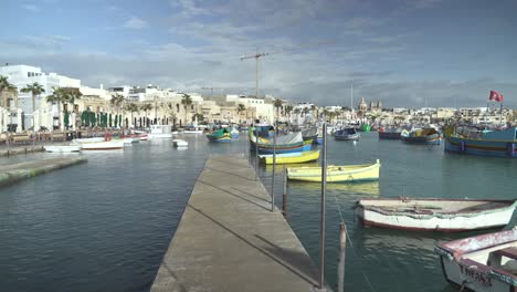 Gehen-Sie-Auf-Einem-Schmalen-Pier-In-Richtung-Traditioneller-Maltesischer-Fischerboote,-Die-Wasser-In-Marsaxlokk-Schaukeln