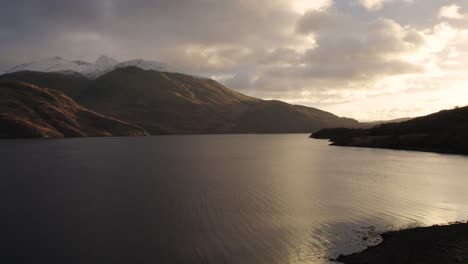 Aerial-drone-footage-slowly-reversing-over-Loch-Etive-and-Glen-Etive-in-the-Highlands-of-Scotland-in-winter-with-snow-capped-mountains-and-a-golden-sunset-reflecting-in-the-water