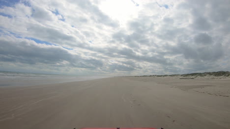 POV-thru-windshield-of-vehicle-driving-on-beach-between-surf-and-dunes-of-North-Padre-Island-National-Seashore-other-vacationers-on-beach-a-cloudy-day