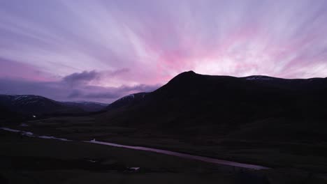 Aerial-drone-footage-of-Glenshee-in-Scotland-during-an-intense-pink-and-purple-sky-changes-colour-looking-towards-the-silhouetted-mountains-as-the-sunset-reflects-in-a-river-and-off-of-the-clouds