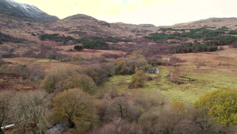 Aerial-drone-footage-slowly-flying-over-trees-in-Glen-Etive-in-the-Scottish-Highlands-towards-Cadderlie-Bothy-in-winter-set-in-a-snow-capped-mountain,-native-broadleaf-woodland-and-a-river-landscape