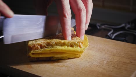 A-close-up-shot-of-the-hands-of-a-chef-positioning-a-sandwich-before-slicing-through-the-middle-with-a-kitchen-knife,-the-cooked-Bacon,-egg-and-cheese-sandwich-is-then-placed-for-presentation