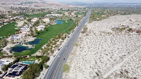 Highway-road-leading-through-green-oasis-town-surrounded-by-vast-deadly-desert,-aerial-drone-shot