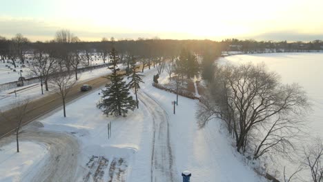 Aerial-view-of-a-park-covered-in-snow-during-a-winter-sunset-with-cars-driving-by,-in-Minneapolis