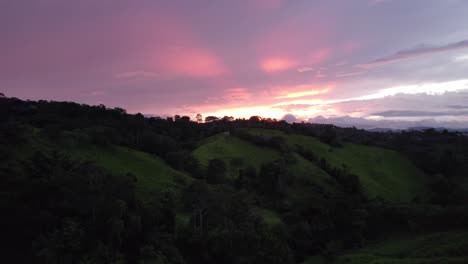 Aerial-dolly-in-flying-over-green-grass-hills-with-dense-forest-on-a-colorful-cloudy-sunset-in-Costa-Rica