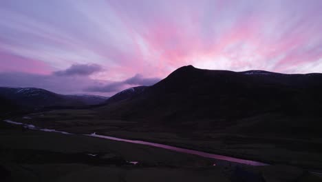 Aerial-drone-footage-rising-slowly-in-Glenshee,-Scotland-during-an-intense-pink-and-purple-sunset-looking-towards-the-silhouetted-mountains-as-the-sunset-reflects-in-a-river-and-off-of-the-clouds