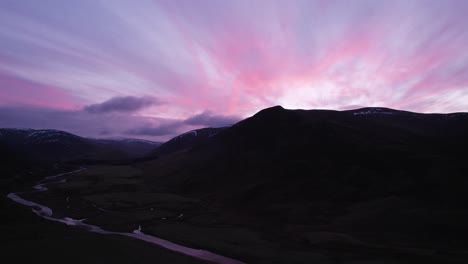 Aerial-drone-footage-of-Glenshee-in-Scotland-during-an-intense-pink-and-purple-sunset-looking-up-the-glen-towards-the-silhouetted-mountains-as-the-sunset-reflects-in-a-river-and-off-of-the-clouds