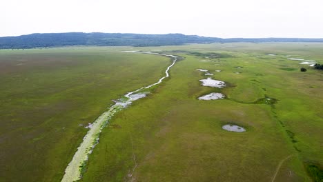 Expansive-green-plain-landscape-with-stream,-river-and-lake-waterways-and-mountains-in-the-background,-aerial-drone-flight-over-wetlands