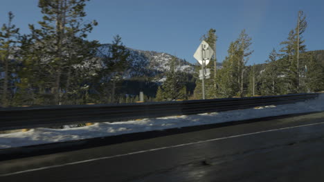 Trees-and-road-signs-pass-by-as-seen-from-the-road-in-front-of-a-mountain-landscape-in-Lake-Tahoe