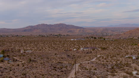 Boom-up-over-dirt-road-and-houses-in-desert-of-Joshua-Tree,-California-with-gorgeous-mountains-on-horizon