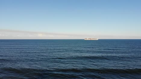 A-large-cargo-ship-sails-through-large,-open-waters-off-the-coast-to-a-nearby-beach-town-under-a-baby-blue-sky-that-harbours-low-hanging-fluffy-clouds