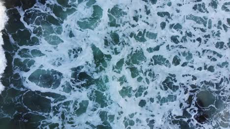 Birds-eye-view-of-a-beautiful-lime-green-and-dark-blue-ocean-as-waves-crash-against-waves-producing-sea-foam-against-dark,-slippering-rocks-as-the-afternoon-sunset-casts-dark,-harsh-shadows