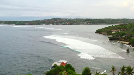 Nusa-Ceningan-Island-coastline-with-waves-and-surfers-in-water,-aerial