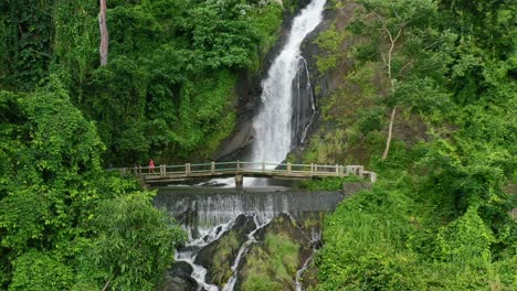 aerial-zoom-out-of-girl-walking-across-a-bridge-at-Kerta-Gangga-Waterfall-in-Lombok