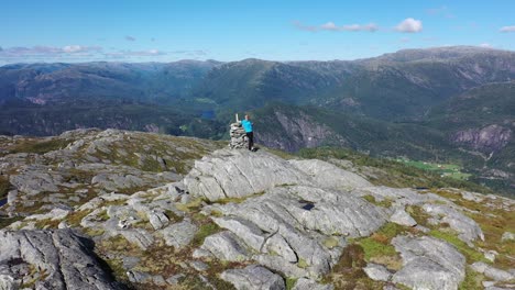 Man-standing-on-Norwegian-mountain-peak-in-sunny-summer-weather---Backward-moving-aerial-from-person-while-revealing-massive-mountain-scenery-in-background---Stamneshella-Vaksdal-Norway